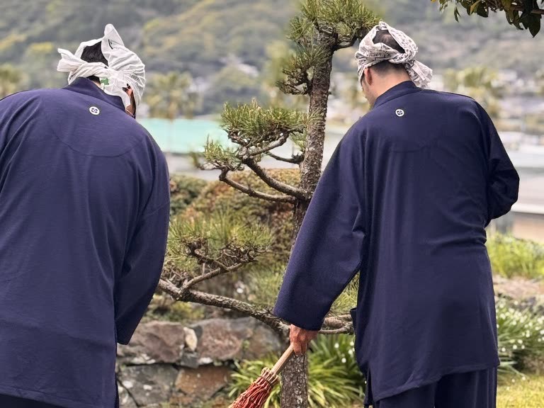 作務衣に手ぬぐいを巻き、竹箒で庭を掃く二人
